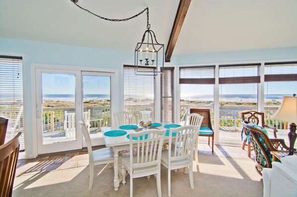 A dining area with a table and chairs, large windows with blinds, and a view of the ocean.