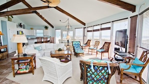 A spacious living room with a high ceiling, wooden beams, and a large window overlooking the ocean. There are several wicker chairs and a white sofa. - Viewpoint, Oceanfront House in Emerald Isle, NC! (Emerald Isle)