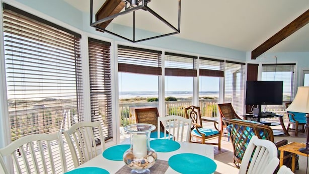 A dining area with a table set for four, surrounded by white chairs with blue cushions. There is a large window with blinds, a television, and a lamp on a side table. - Viewpoint, Oceanfront House in Emerald Isle, NC! (Emerald Isle)