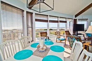 A dining area with a table set for four, surrounded by white chairs with blue cushions. There is a large window with blinds, a television, and a lamp on a side table. - Viewpoint, Oceanfront House in Emerald Isle, NC! (Emerald Isle)