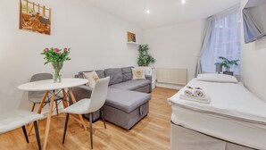 A modern living room with a grey sofa, a round dining table with chairs, a small white table with a vase of flowers, and a bed with towels. - Central London apartment (London)