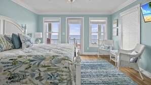 A bedroom with a large bed, a ceiling fan, and a view of the ocean through multiple windows. - Blue View, Oceanfront House in Pine Knoll Shores, NC! (Pine Knoll Shores)