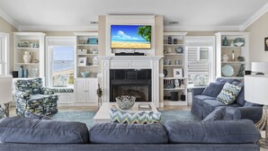 A living room with a fireplace, a television mounted above it, and a view of the beach through the windows. - Blue View, Oceanfront House in Pine Knoll Shores, NC! (Pine Knoll Shores)