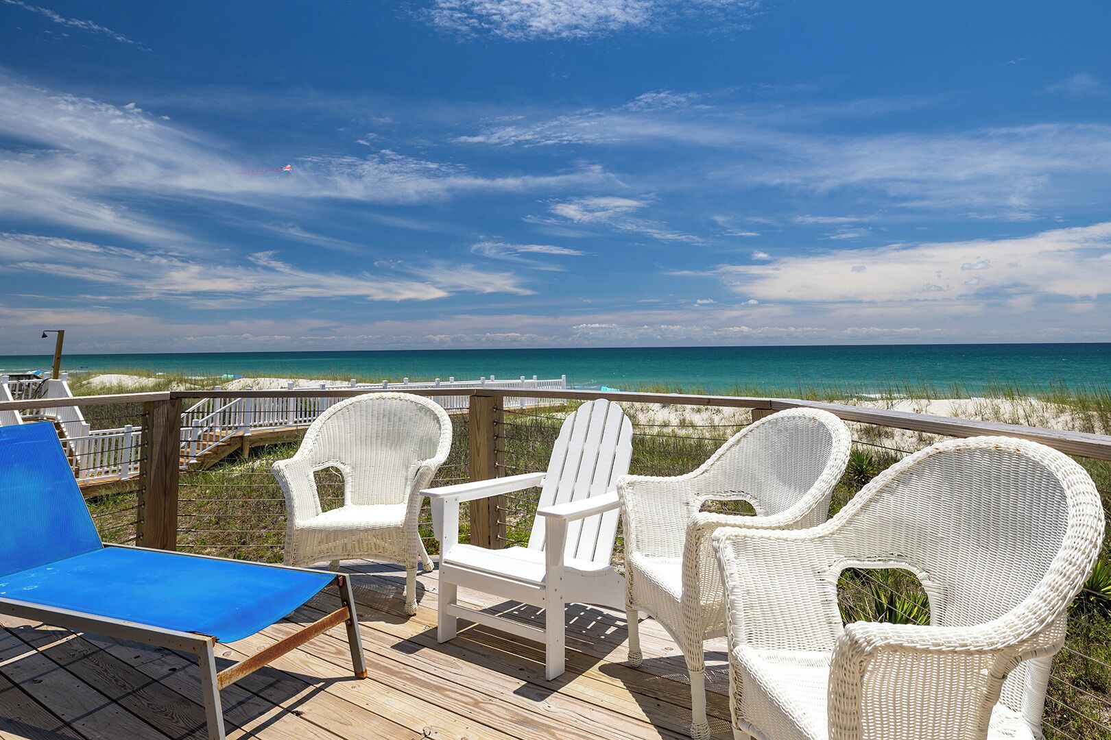 A wooden deck with white wicker chairs and a blue lounge chair overlooking a beach.