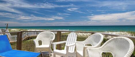 A wooden deck with white wicker chairs and a blue lounge chair overlooking a beach.