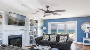 Ein Wohnzimmer mit Kamin, großem Fernseher, Deckenventilator und Meerblick durch ein großes Fenster.