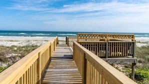 Een houten looppad naar een strand, met een houten omheining en een gebouw rechts.