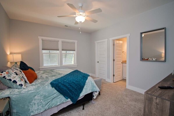 Um quarto com cama, ventilador de teto, janela com persianas, porta e espelho na parede.