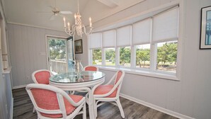 Un coin repas avec une table en verre et des chaises en osier blanc, de grandes fenêtres équipées de stores et un lustre.