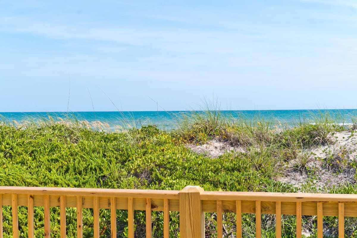 A wooden deck overlooking a beach with green vegetation and the ocean in the background.