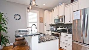 A modern kitchen with white cabinets, a stainless steel refrigerator, a built-in oven, and a granite countertop. There are wooden stools and a potted plant. - Bungalows At The Grove #6, Oceanside House in Atlantic Beach, NC! (Atlantic Beach)