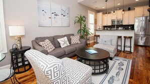 A modern living room with a grey sofa, patterned armchairs, a round coffee table, and a dining area with bar stools. A kitchen with stainless steel appliances is visible in the background. - Bungalows At The Grove #6, Oceanside House in Atlantic Beach, NC! (Atlantic Beach)