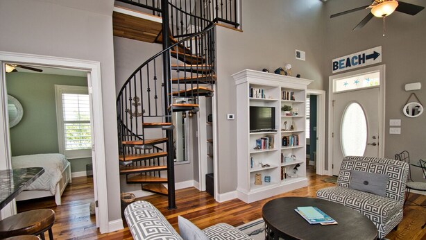 A living room with a spiral staircase, a bookshelf, a sofa, and a coffee table. - Bungalows At The Grove #6, Oceanside House in Atlantic Beach, NC! (Atlantic Beach)