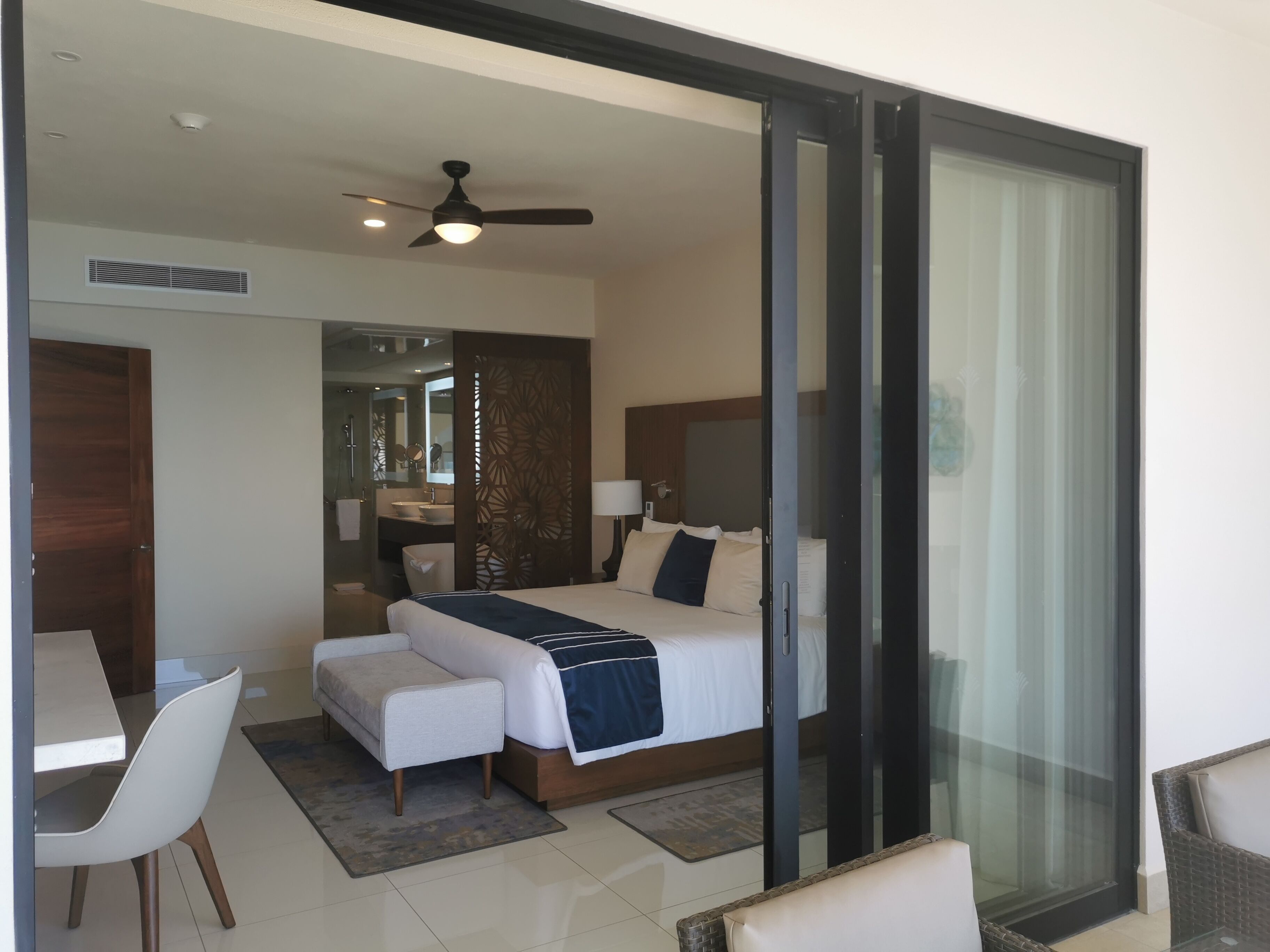 A modern hotel room with a bed, a desk, a chair, a sofa, and a glass door leading to a balcony.