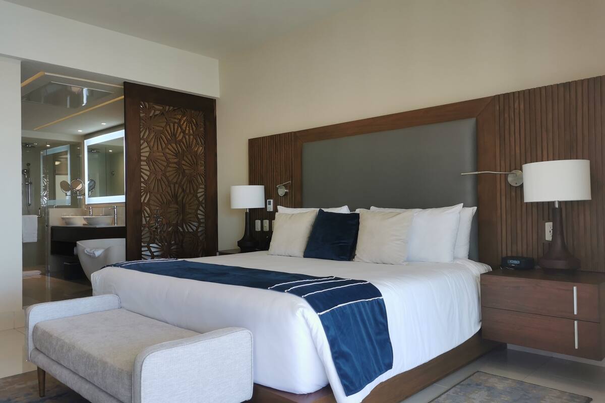 Ein modernes Hotelzimmer mit einem großen Bett, Nachttischlampen, einem Holz-Kopfteil, einem kleinen Nachttisch und einer Sitzecke mit einem Sessel.