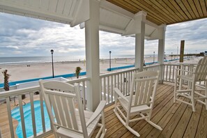 Une terrasse en bois avec des fauteuils à bascule blancs, donnant sur une plage et une piscine.