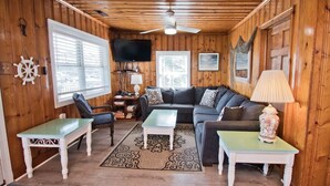 A cozy cabin interior with a wooden ceiling, a sofa set, two tables, a television, and a ceiling fan. - Captain's Escape, Almost Oceanfront House in Emerald Isle, NC! (Emerald Isle)