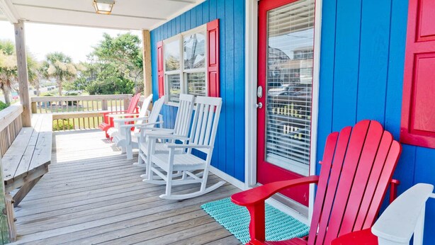 A wooden deck with white rocking chairs and a red chair, a red door, and a window with blinds. - Captain's Escape, Almost Oceanfront House in Emerald Isle, NC! (Emerald Isle)
