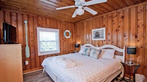 A wooden-paneled bedroom with a bed, a ceiling fan, a window, a television, and a small table. - Captain's Escape, Almost Oceanfront House in Emerald Isle, NC! (Emerald Isle)