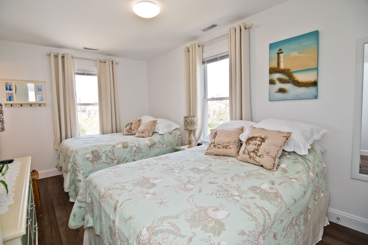 A bedroom with two beds, a painting of a lighthouse, and a window with curtains.