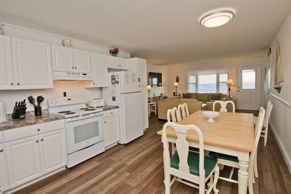 A kitchen with white cabinets, a wooden dining table with chairs, and a view of the sea through the windows.