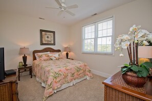 A bedroom with a bed, two bedside tables, a ceiling fan, a window with blinds, and a wall-mounted picture. - Carolina, Oceanside House in Emerald Isle, NC! (Emerald Isle)