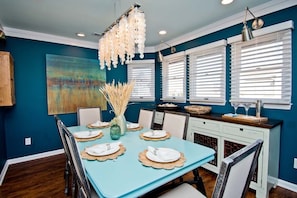 A dining area with a blue table set for four, white chairs, a chandelier, and a large abstract painting on the wall.
