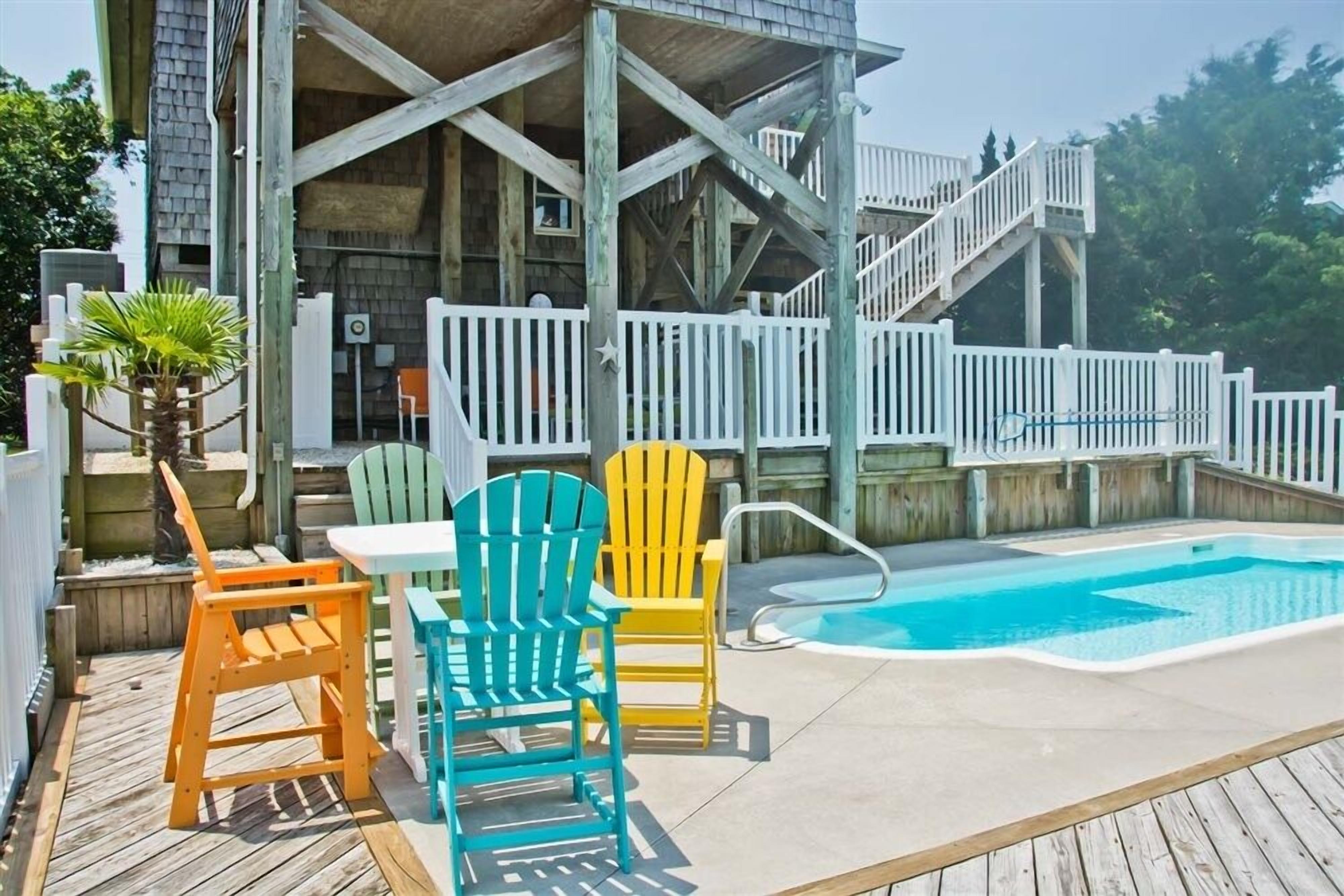 A wooden deck with a pool, Adirondack chairs, and a white fence.