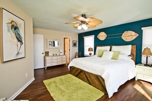 A bedroom with a wooden headboard, a green rug, a ceiling fan, and a wall with a bird painting. - Carolina Conch House, Almost Oceanfront House in Emerald Isle, NC! (Emerald Isle)
