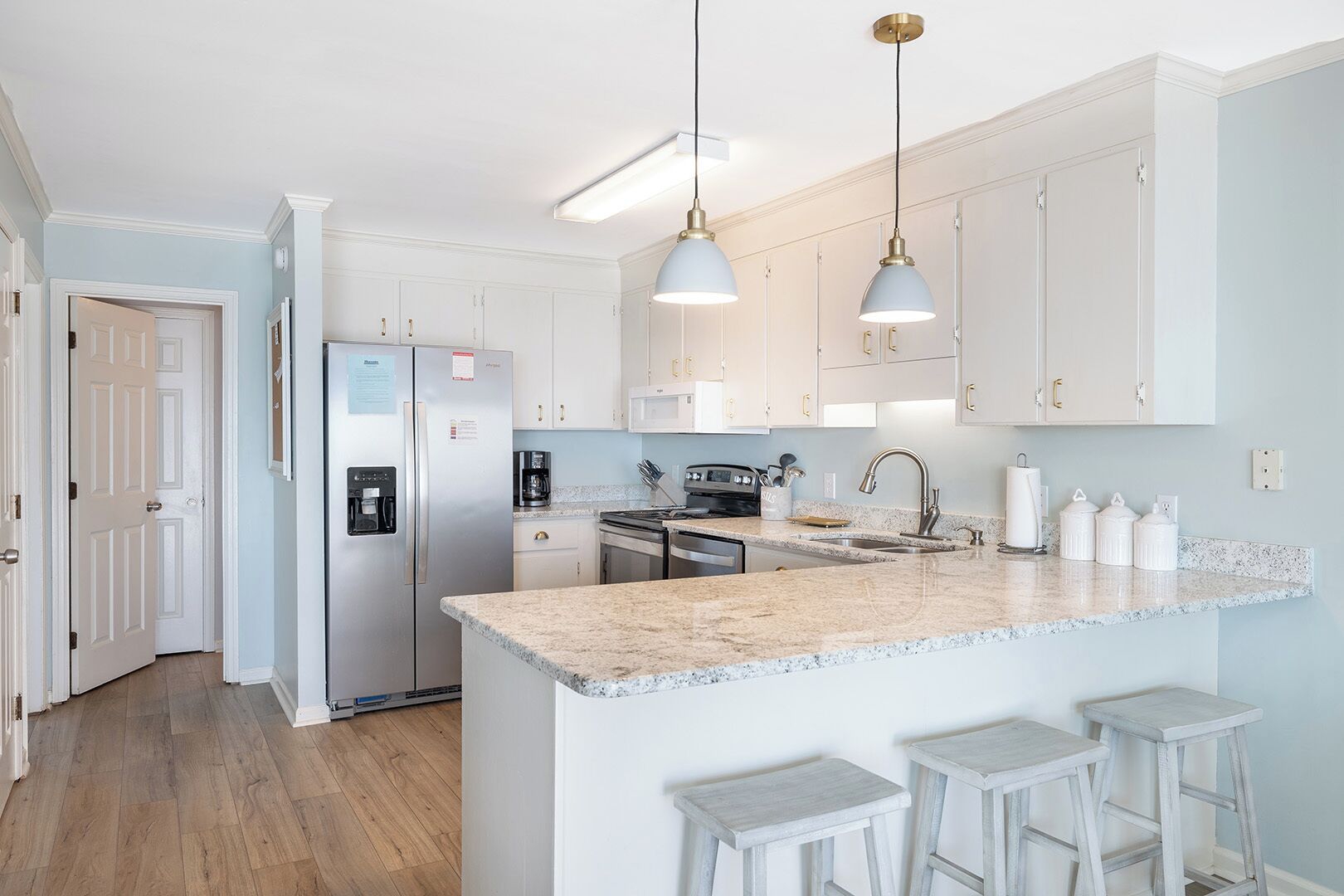 Eine moderne Küche mit weißen Schränken, einer Granit Arbeitsplatte und einem eingebauten Backofen. Zwei Pendelleuchten, ein Kühlschrank und eine Kaffeemaschine sind vorhanden.
