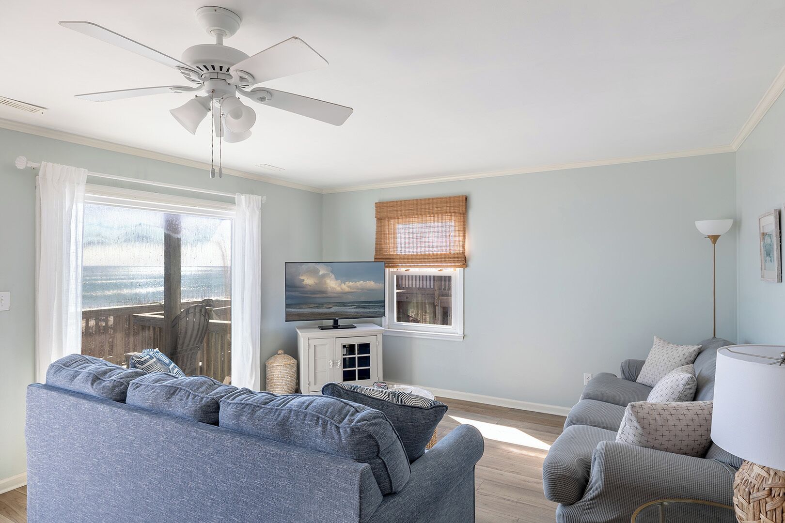 Ein Wohnzimmer mit Deckenventilator, Fernseher auf einem Schrank und Meerblick durch ein großes Fenster.