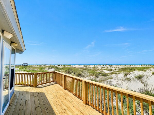 Une terrasse en bois avec une porte vitrée, donnant sur une plage bordée de dunes et l’océan.