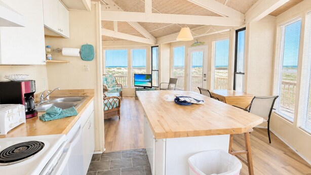 Une cuisine avec un îlot central en bois, un coin repas avec des chaises, un téléviseur et une vue sur la plage.