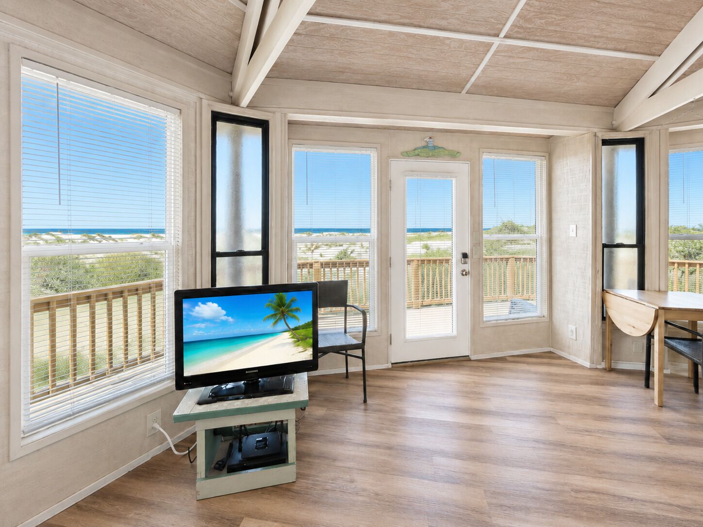 A room with a television on a stand, a chair, and a view of the beach through large windows.