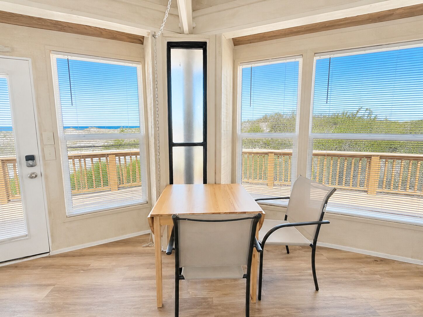 A room with a wooden table and two chairs, large windows with blinds, and a view of the outdoors.