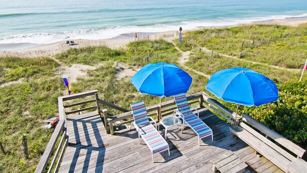 A wooden deck with striped lounge chairs and blue umbrellas overlooking a beach. - Casa Atlantica, Oceanfront House in Pine Knoll Shores, NC! (Pine Knoll Shores)