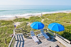 A wooden deck with striped lounge chairs and blue umbrellas overlooking a beach. - Casa Atlantica, Oceanfront House in Pine Knoll Shores, NC! (Pine Knoll Shores)