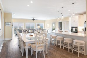 A modern kitchen with white cabinetry, a central island with stools, and a dining area with a white table and chairs. - Casa Del Sole, Oceanfront House in Emerald Isle, NC! (Emerald Isle)
