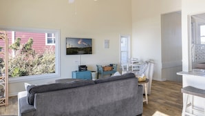 Ein Wohnzimmer mit grauem Sofa, einem an der Wand befestigten Fernseher, einem Deckenventilator und Blick auf ein rotes Gebäude.
