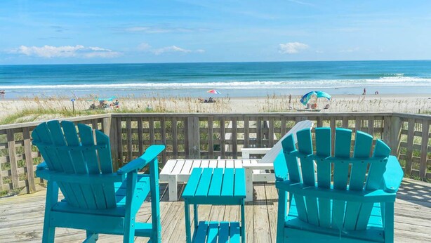 Une terrasse en bois, agrémentée de chaises Adirondack turquoise et d’une petite table, donnant sur une plage où l’on aperçoit des gens et des parasols.
