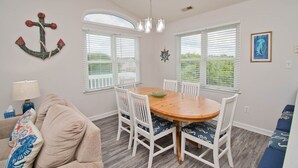 A dining area with a wooden table and white chairs, large windows with blinds, a chandelier, and a wall anchor decoration. - Chillin' The Most, Oceanview House in Emerald Isle, NC! (Emerald Isle)