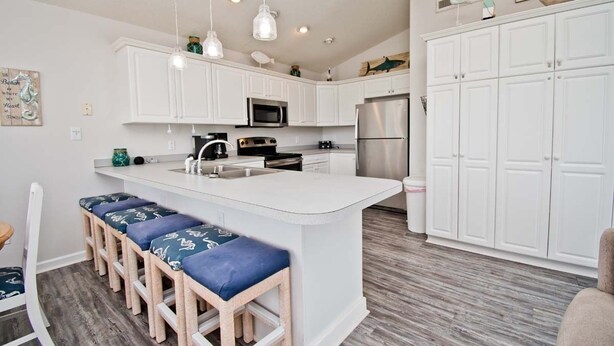 A modern kitchen with white cabinetry, a central island with bar stools, stainless steel appliances, and pendant lighting. - Chillin' The Most, Oceanview House in Emerald Isle, NC! (Emerald Isle)