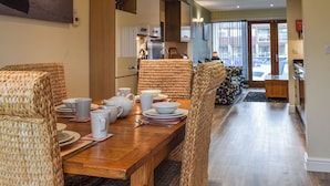 A dining area with a wooden table set for four, wicker chairs, and a kitchen in the background. - 2 bedroom accommodation in Carnforth (Carnforth)
