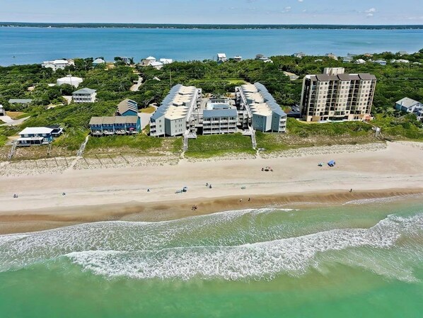 An aerial view of a coastal hotel with multiple buildings, a sandy beach, and a clear view of the ocean.