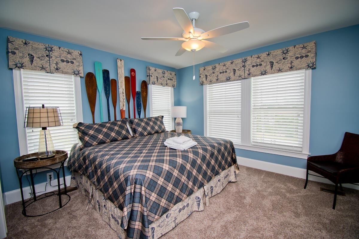 A bedroom with a bed, two nightstands, a chair, a ceiling fan, and windows with shutters.