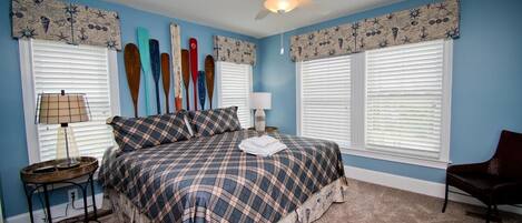 A bedroom with a bed, two nightstands, a chair, a ceiling fan, and windows with shutters.