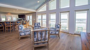 A spacious room with large windows, a wooden floor, and wicker furniture. - Compass Pointe, Oceanfront House in Emerald Isle, NC! (Emerald Isle)