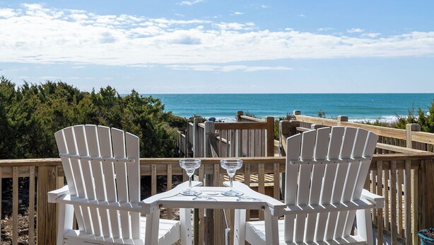 Een houten terras met twee witte Adirondack-stoelen en een tafel, met uitzicht op een strand, de oceaan en een heldere lucht.