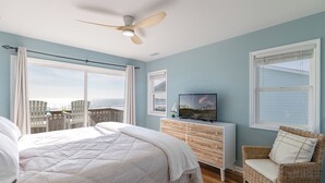 A bedroom with a bed, a dresser, a television, a wicker chair, and a view of the ocean. - Angel's at the Pier West - Oceanfront Duplex in Emerald Isle, NC! (Emerald Isle)
