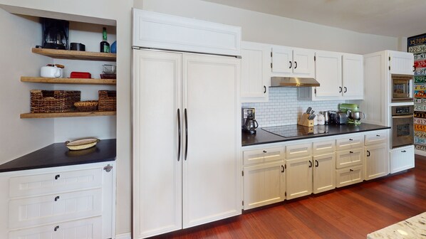 Una cocina moderna con armarios blancos, encimera negra y campana extractora de acero inoxidable. Se observan estantes de madera con cestas y utensilios de cocina.